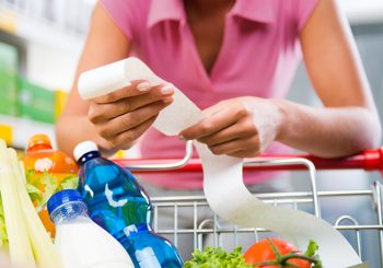 Woman in pink polo shirt leans over grocery cart containing vegetables and several bottled beverages and reviews a long receipt.