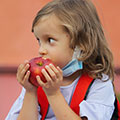 young child wearing backpack, mask pulled below chin, eating apple