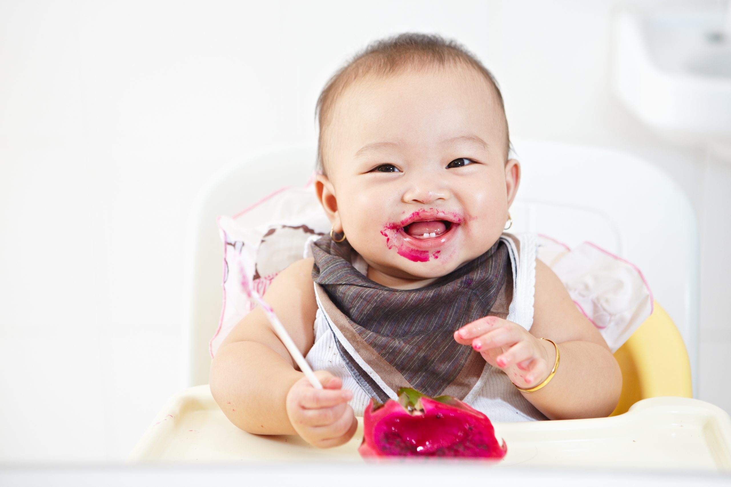 Baby in high chair eating red dragon fruit, smiling